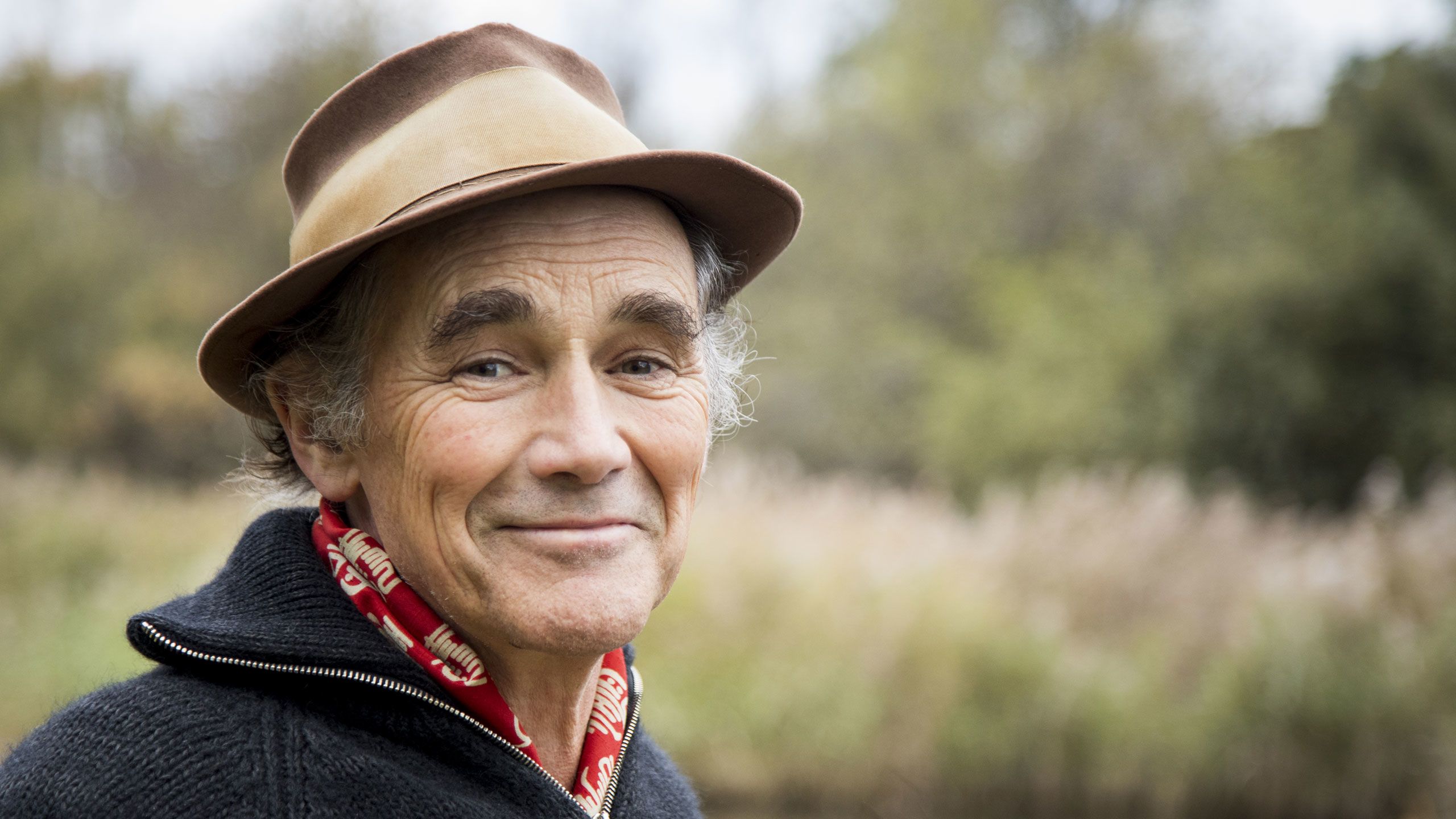 New WWT ambassador Sir Mark Rylance smiling at the camera, with a wetland backdrop